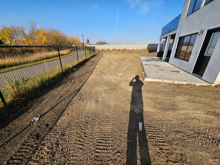 Long shadow of person on dirt ground beside metal fence and modern building under clear sky