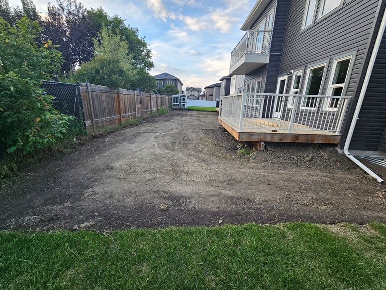 Residential backyard with dirt area, green lawn, house with deck, wooden fence, and trees under cloudy sky