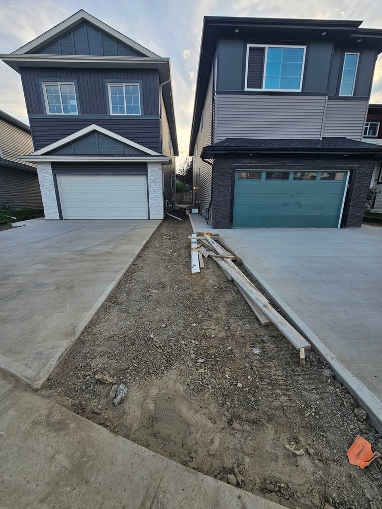 Two neighboring two-story homes with dark siding and garage doors separated by a narrow dirt pathway under construction