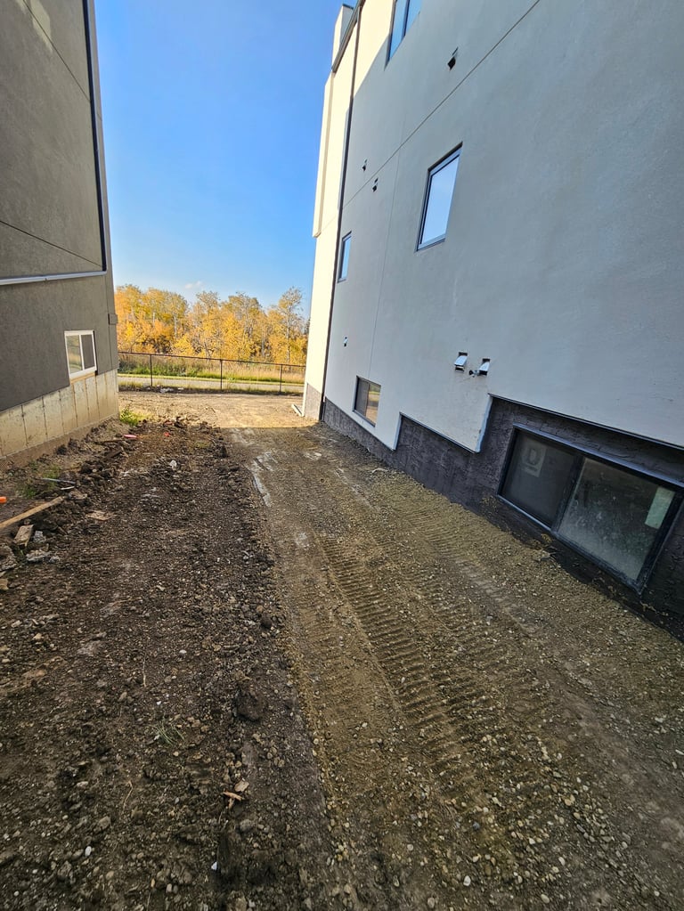 Narrow side alley between two modern white buildings with gravel ground and autumn trees visible in the distance