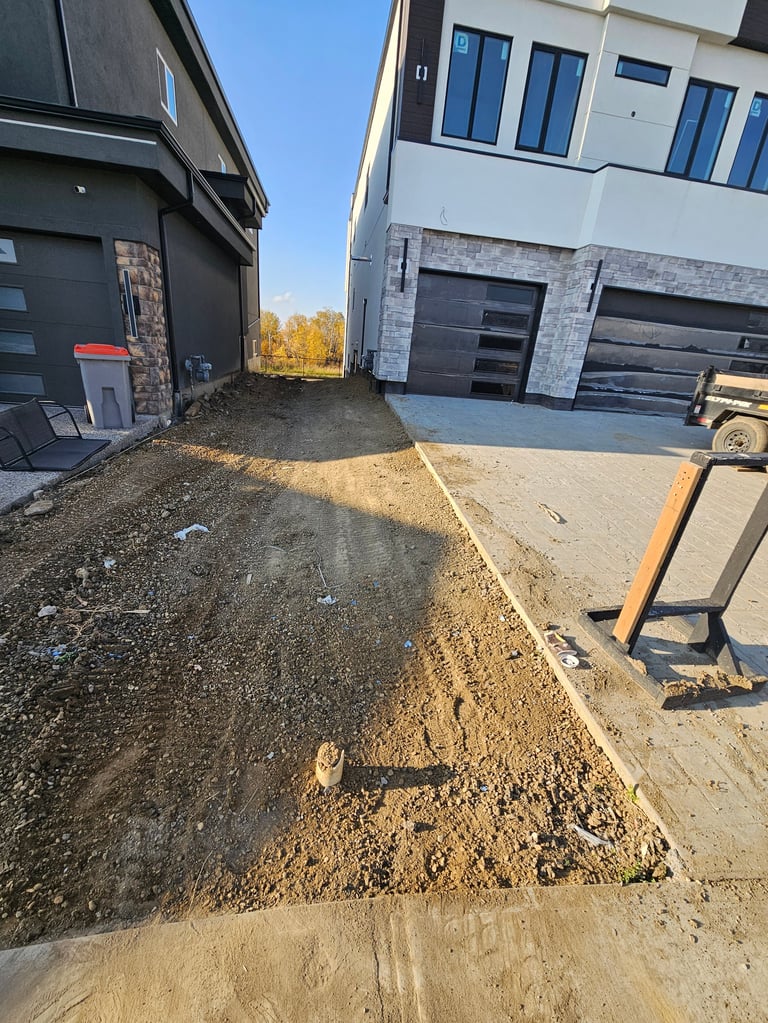 Construction site between two modern homes showing dirt driveway, concrete foundation, and garage doors under development