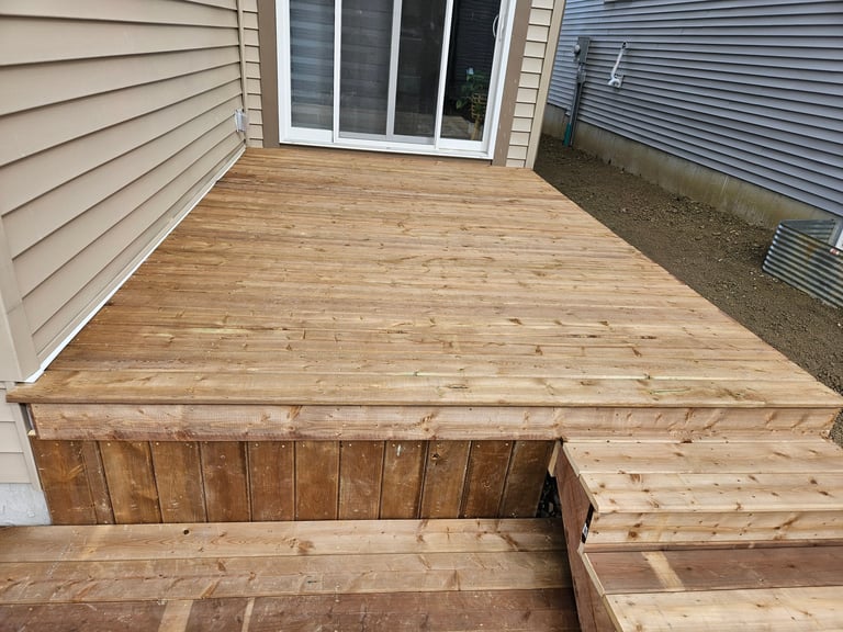 Newly built wooden deck with light brown stained boards attached to house with white siding and glass door